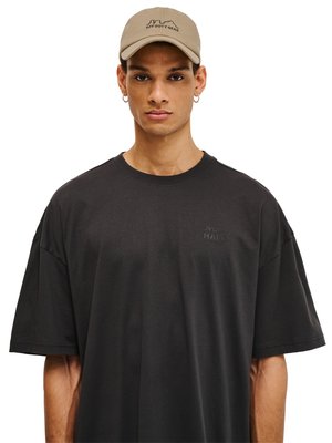Jonge man met een beige pet met het logo "OFF DUTY GEAR" en een zwart oversized T-shirt met een subtiel "HALO" logo op de borst, kijkt recht vooruit.