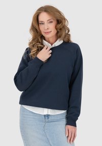 Felpa blu navy con collo rotondo, maniche raglan e polsini a costine, indossata sopra una camicia bianca con colletto. Tessuto morbido, vestibilità casual.
