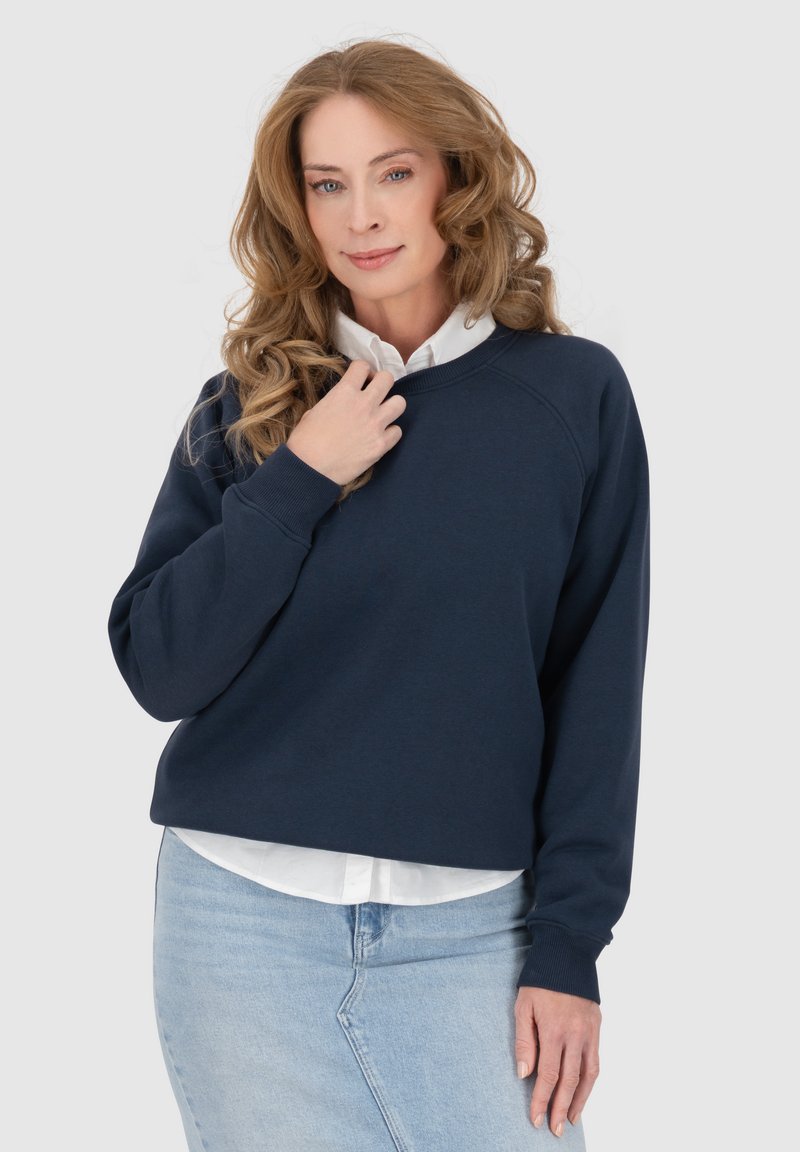 Felpa blu navy con collo rotondo, maniche raglan e polsini a costine, indossata sopra una camicia bianca con colletto. Tessuto morbido, vestibilità casual.