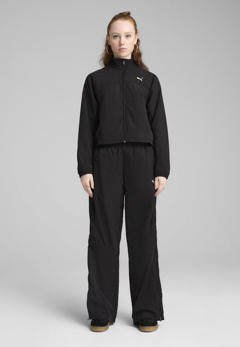 Črnen športni komplet, ki vključuje skrajšano jakno na zadrgo in ohlapne hlače, izdelan iz lahkega materiala, z diskretnim logotipom na obeh kosih.