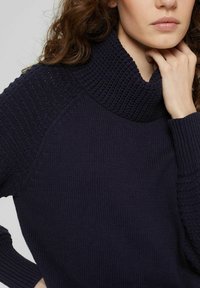 Pull en tricot bleu marine avec un col haut texturé et des manches côtelées. Présente un mélange de motifs lisses et tissés.