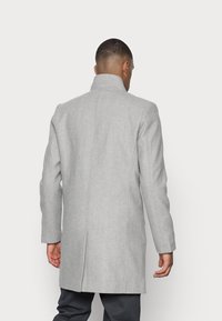 Uomo di spalle che indossa un cappotto grigio chiaro fino al ginocchio con colletto alto e pantaloni scuri su uno sfondo neutro.