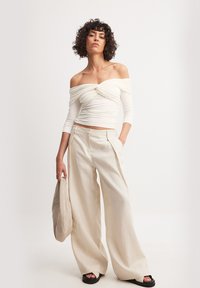 Off-the-shoulder witte top met gathered detail, gecombineerd met losse beige linnen broek. Model houdt een bijpassende beige tas vast en draagt zwarte sandalen.