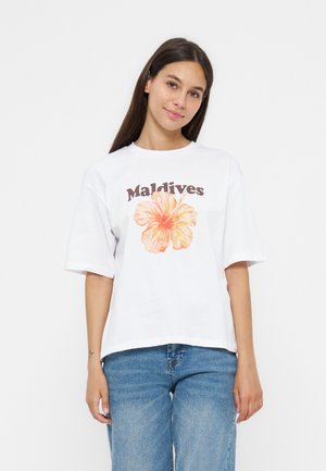 Jeune femme aux longs cheveux foncés portant un t-shirt blanc avec un grand hibiscus orange et le texte "Maldives", associé à un jean bleu.
