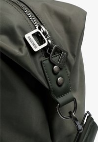 Mochila de tela en verde oliva con un zipper plateado, detalles de cuero y accesorios de metal. Incluye una correa con botones a presión.
