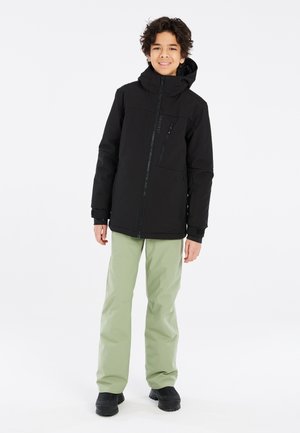 Schwarze wasserdichte Jacke mit hohem Kragen, Frontreißverschluss und Logodetail, kombiniert mit hellgrünen Hosen und schwarzen wasserdichten Stiefeln.