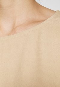 Beige topp i en slät, texturerad tyg. Har en bred, rund halsringning och fina sömmar längs kanten.