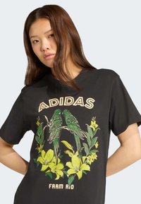 Juodas medvilnės marškinėlis su dviem žaliais paukščiais ir geltonomis gėlėmis, virš dizaino esantis kremiškas tekstas "ADIDAS" ir "FARM RIO".