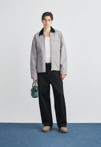 Γκρι ζακέτα με φερμουάρ και μαύρο γιακά, λευκό πουκάμισο από κάτω, μαύρο παντελόνι φαρδύ στο κάτω μέρος και πράσινη τσάντα χειρός. Διαθέτει έμβλημα με λογότυπο στη ζακέτα.