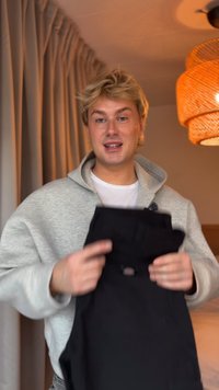 Crna čvrsta kecelja s prednjim džepom i prilagodljivim trakama, koju drži osoba u svijetlosivoj dukserici na neutralnoj pozadini.