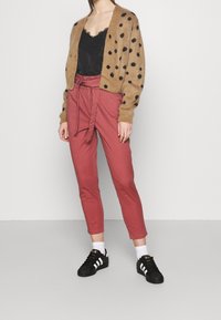 Bruine polkadot cardigan over een zwarte kanten top, gecombineerd met tapered rode broek en zwarte sneakers met witte strepen. Casual outfit.