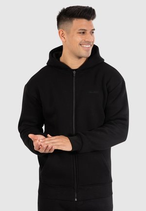 Sort zip-up hoodie lavet af blødt stof. Den har en hætte med snoretræk, frontlommer, ribbede manchetter og en simpel logo detalje på brystet.
