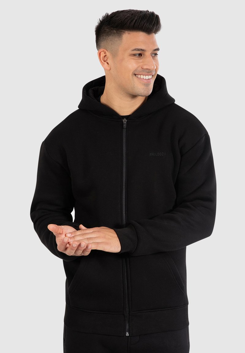 Zwarte zip-up hoodie van zacht materiaal. Het heeft een capuchon met trekkoord, voorzakken, ribgebreide manchetten en een eenvoudig logo op de borst.