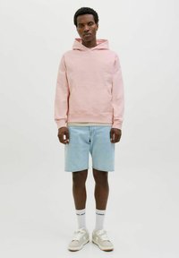 Lichtroze hoodie met een kangoeroezak, relaxed fit, gecombineerd met lichtdenim shorts en beige sneakers. Witte sokken met zwarte tekst.
