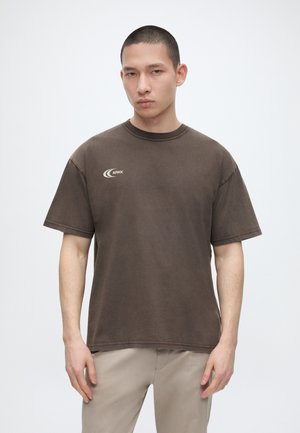 Młody mężczyzna z krótkimi włosami, noszący brązową koszulkę z logo ARKK i beżowe spodnie, stoi na jednolitym białym tle.