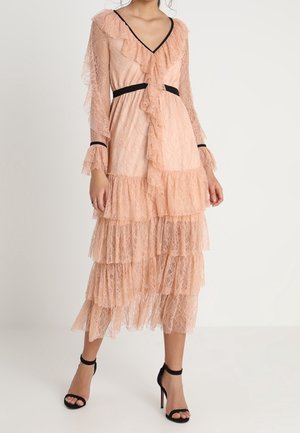 Kobieta ubrana w wielowarstwową, brzoskwiniową sukienkę midi z koronki z czarnymi wykończeniami w talii i na mankietach, z dekoltem w serek, falbankami oraz czarne sandały na wysokim obcasie.