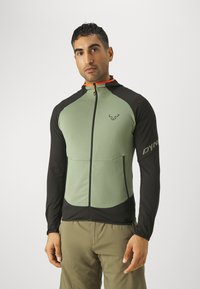 Green and black jacket with long sleeves, zipper, and logo on sleeve. Features contrasting colors and a fitted design, made from breathable material.
