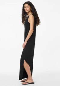 Juoda maksi suknelė su briaunotu paviršiumi, be rankovių, šoniniu įpjovimu ir laisvu siluetu. Apsirengta su juodais šlepetėmis.