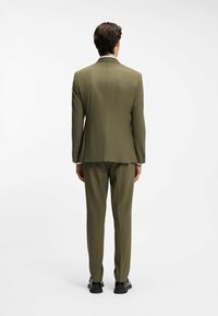 Traje verde oliva con blazer ajustado y pantalones rectos. Diseño minimalista, tela texturizada y zapatos negros que complementan el conjunto.