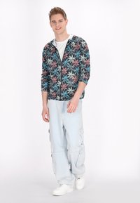 Jachetă cu glugă și fermoar, cu un model colorat de frunze pe un fundație neagră; asortată cu pantaloni cargo de culoare bleu deschis și adidași albi.
