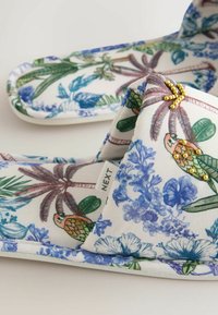 Šlepetės su paminkšlintomis juostelėmis, puoštos tropinių gėlių ir papūgos raštu, dekoruotos mažais aukso karoliukais, šviesiame fone.