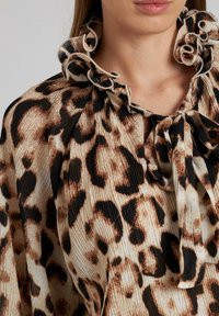Blusa de estampado de leopardo con cuello con volantes, con patrones marrones y negros sobre un fondo crema, hecha de una tela ligera plisada.