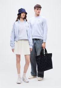 Γυναίκα με γαλάζιο μπουφάν, λευκή plisέρ φούστα, αθλητικά παπούτσια και σκούφο τύπου bucket σε ναυτικό μπλε χρώμα στέκεται δίπλα σε άνδρα με γαλάζια μπλούζα, τζιν, λευκά αθλητικά παπούτσια, που κρατά μαύρη τσάντα tote.