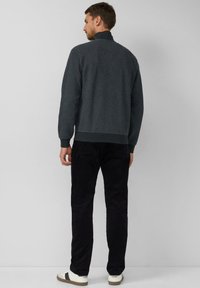 Dunkelgrauer strukturierten Pullover mit hohem Kragen, getragen über schwarzen Cordhosen und weißen Sneakers mit schwarzen Akzenten. Ansicht von hinten.