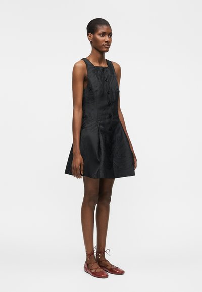 Γυναίκα όρθια, φοράει μαύρο αμάνικο φόρεμα με κουμπιά και κόκκινα δερμάτινα παπούτσια με κορδόνια, με ουδέτερη έκφραση προσώπου σε απλό φόντο.