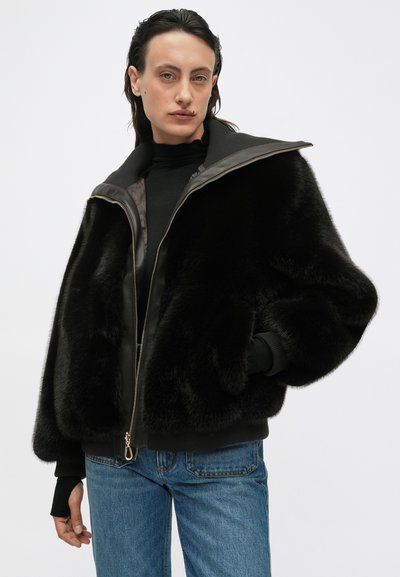 Giacca nera in pelliccia ecologica con zip frontale, ampio colletto e polsini a coste. Presenta un rivestimento liscio e un design minimalista, indossata con jeans blu.