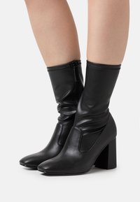 Botas de tobillo de imitación cuero negro con un diseño arrugado, punta afilada y un tacón grueso. Presenta una cremallera lateral para un fácil uso.