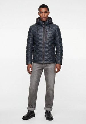 Mann trägt eine dunkle wattierte Kapuzenjacke, graue hochgekrempelte Jeans und schwarze Stiefel, steht vor einem schlichten weißen Hintergrund.