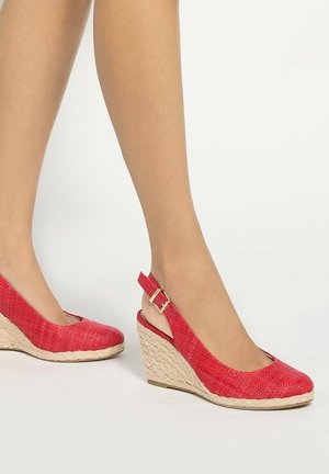 Feet wearing red textured slingback wedge shoes with braided rope soles on a white background.