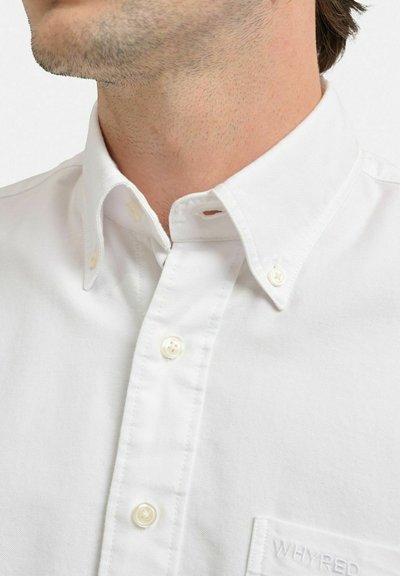Vit bomullsskjorta med knapp-ner krage, synliga vita knappar, vänster ficka, och subtilt broderad logotyp på fickan.