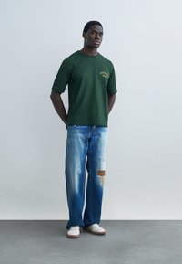 Jaunuolis stovintis rankomis už nugaros, vilkintis tamsiai žalią trumpomis rankovėmis marškinius, taisytus mėlynus džinsus ir dėvintis baltus sportinius batelius ant pilkos grindų.