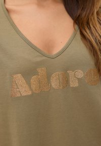 T-shirt vert olive à col V avec le texte "Adore" en doré pailleté ; tissu doux, coupe décontractée, design minimal, sans motifs ni accents au-delà du texte.
