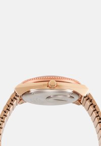 Montre-bracelet en or rose avec une lunette texturée, un boîtier rond et un bracelet en maille métallique. Comprend une couronne proéminente et un fond en acier inoxydable.