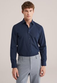 Jonge man met krullend haar, gekleed in een marineblauw overhemd met knopen, ingestopt in lichtgrijze broek, staand tegen een effen achtergrond.