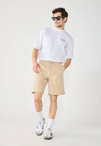 White t-shirt with logo, beige shorts, white socks, and black and white sneakers. Model poses with hands in pockets, wearing dark sunglasses.