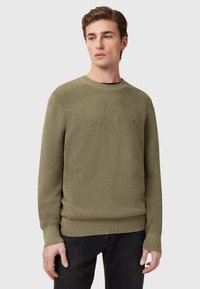 Khaki trui van gemêleerd breimateriaal, met een ronde hals en geribbelde manchetten, gecombineerd met donkere spijkerbroek.