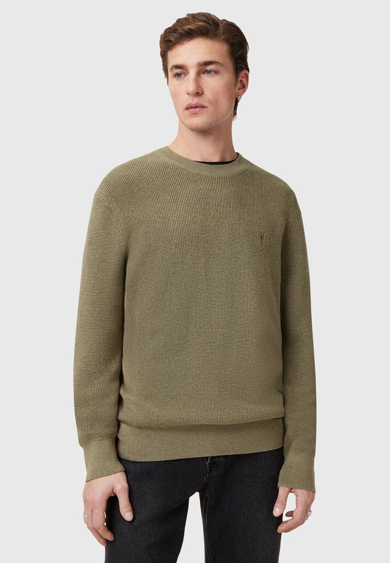 Khaki trui van gemêleerd breimateriaal, met een ronde hals en geribbelde manchetten, gecombineerd met donkere spijkerbroek.