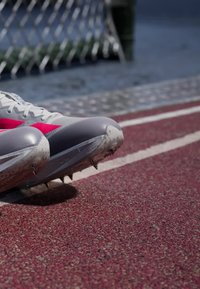 Grå og lyserøde pigge til løbesko placeret på en rød løbebane nær et metalhegn med kæder.