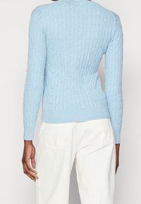 Osoba w jasnoniebieskim swetrze w splot z kabli i białych spodniach, pokazana z tyłu, stojąca na jednolitym tle.