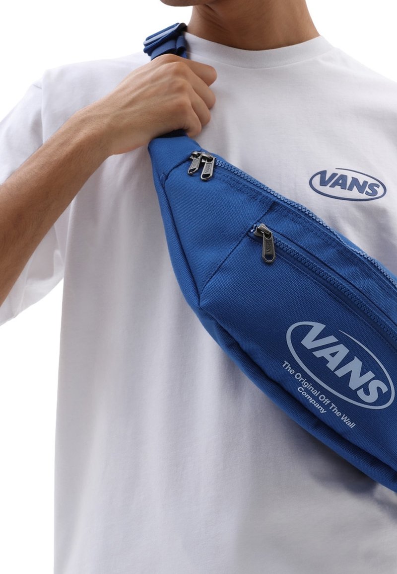 Personne portant un t-shirt blanc Vans tenant un sac bandoulière bleu Vans avec des fermetures éclair et logos visibles.