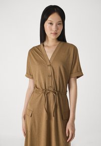 Rochie maro, cu mânecă scurtă și decolteu în V, cu închidere prin nasturi și cordon la talie. Prezintă două buzunare frontale și un material moale, texturat.