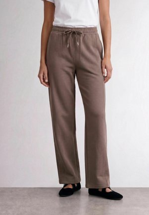 Femme portant un pantalon de survêtement marron à jambes larges avec une taille à cordon et des chaussures plates noires, debout sur un sol gris contre un mur clair.
