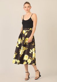 Top nero senza maniche abbinato a una gonna midi floreale marrone con fiori gialli, vita aderente e tasche laterali. Tacchi neri con cinturini.