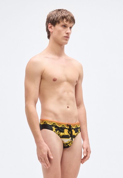 Μαύρα μαγιό σλιπ με χρυσό μπαρόκ σχέδιο, πορτοκαλί ζωνάρι στη μέση και κορδόνι για ρύθμιση. Η υφή του υφάσματος φαίνεται λεία και γυαλιστερή.