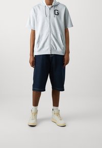 Haut à zip gris avec des manches courtes et des poches avant, associé à un short en jean bleu et des baskets montantes blanches.