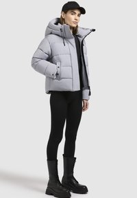 Hellegraue Daunenjacke mit Kapuze, in lässigem Schnitt und genähten Bahnen. Getragen über einem schwarzen Oberteil und schwarzen Leggings, mit geschnürten schwarzen Stiefeln.